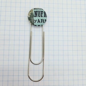 May include: A silver paper clip with a circular fabric button. The button has a light blue background and black text reading "MONSIEUR NUFA D'ARM". The clip is set against a white grid background.