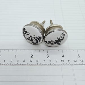 May include: Two round white ceramic cabinet knobs featuring black butterfly illustrations. Each knob has a silver-toned metal base and a threaded screw. A ruler placed in front shows the knobs are approximately 3.8 cm in diameter.