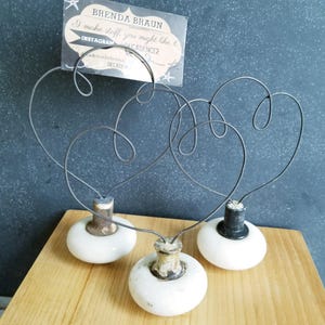 Peut inclure: Trois boutons de porte en céramique blanche avec des accents noirs, chacun surmonté d'un cadre en fil de fer en forme de cœur. Une carte avec le texte "BRENDA BRAUN I make stuff you might like x INSTAGRAM #CADENCE2 decadence2hotmail.com #DECADENCE2" est affichée dans le cadre.