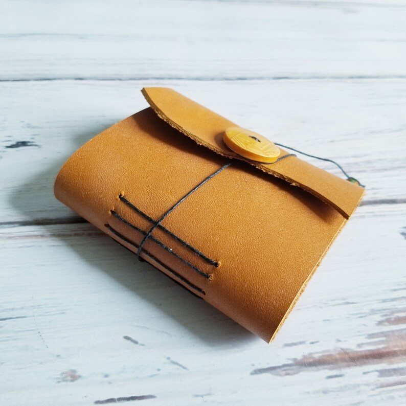 May include: A brown leather journal with a flap closure and a wooden button. The journal is tied closed with a string and has black stitching.