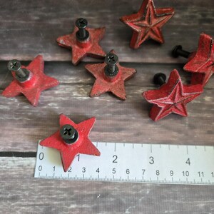 May include: Six red metal star-shaped drawer pulls with black screws. The pulls are arranged on a wooden surface with a ruler for scale.