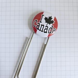 May include: A circular badge with a red and white design, displaying the words "I Canada" and a black maple leaf. The badge is held by a silver paperclip, positioned on a white, grid-patterned surface.