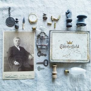 May include: A vintage photograph of a man in a suit and tie, a light bulb, a metal anchor, a small wooden box with the text "Chesterfield" on the lid, a watch, a chess piece, a spool of thread, and other assorted items.