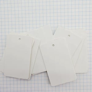 May include: A collection of blank, rectangular white tags with a small hole at the top for stringing. The tags are stacked and scattered, ready for use. The background is a grid pattern.