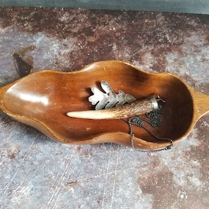 May include: A silver oak leaf pendant necklace with a black chain. The pendant is attached to a piece of antler with a silver cap. The necklace is displayed in a wooden bowl.
