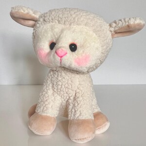 May include: A white plush lamb toy with a pink nose and blush on its cheeks. The lamb is sitting with its legs tucked under its body.