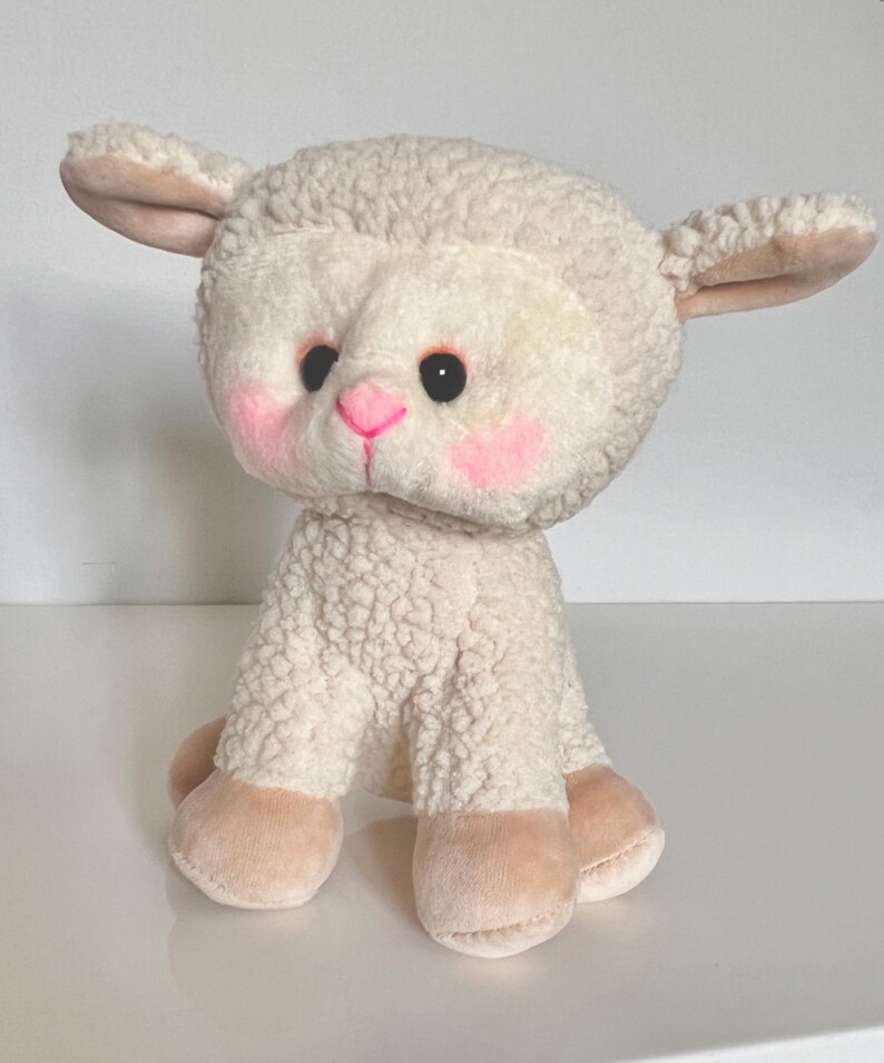 May include: A white plush lamb toy with a pink nose and blush on its cheeks. The lamb is sitting with its legs tucked under its body.
