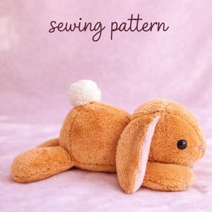 May include: A plush, brown bunny toy with a white pom-pom tail and pink inner ears. The bunny is lying down on a pink surface. The words "sewing pattern" are written in a cursive font above the bunny.
