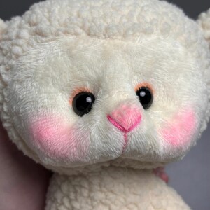 May include: A white plush lamb with pink cheeks and a pink nose. The lamb has black eyes and a fluffy white coat.