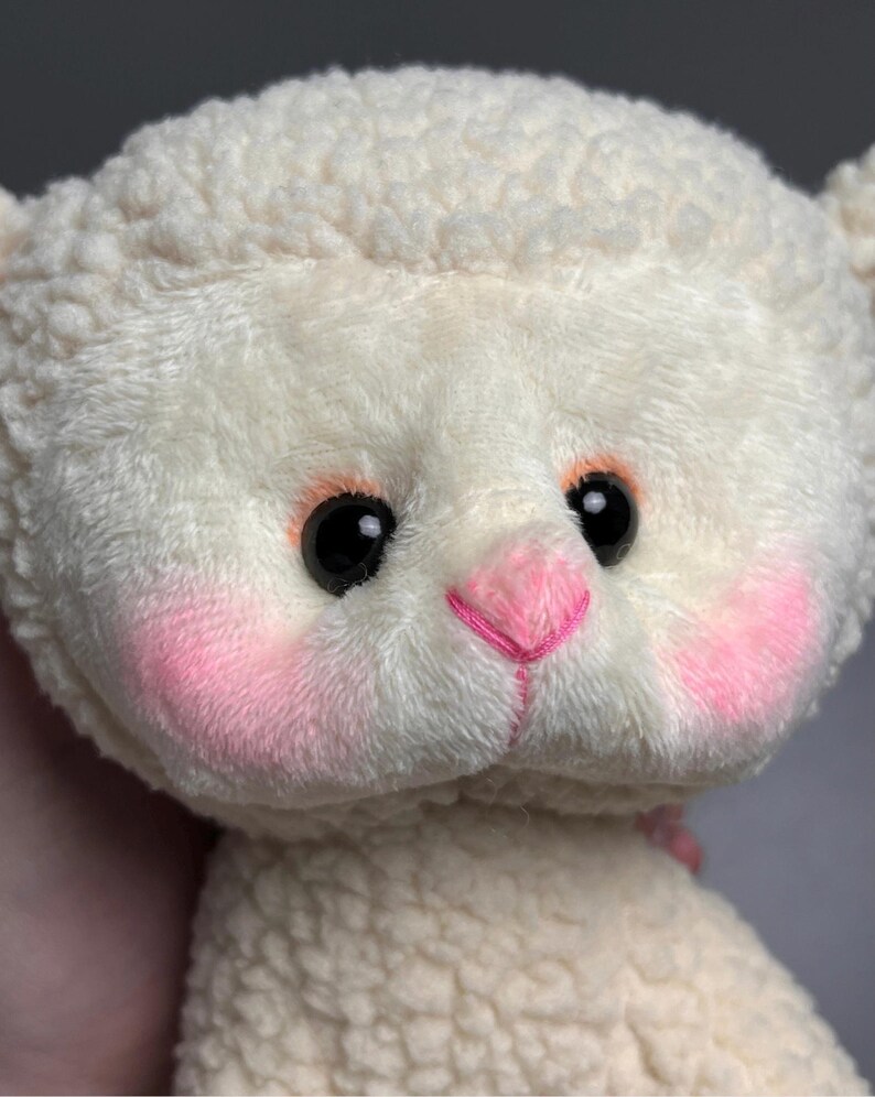 May include: A white plush lamb with pink cheeks and a pink nose. The lamb has black eyes and a fluffy white coat.