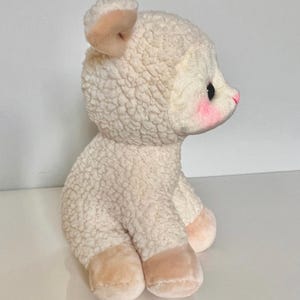 May include: A white plush lamb with a pink nose and blush on its cheeks. The lamb is sitting with its legs tucked under its body.