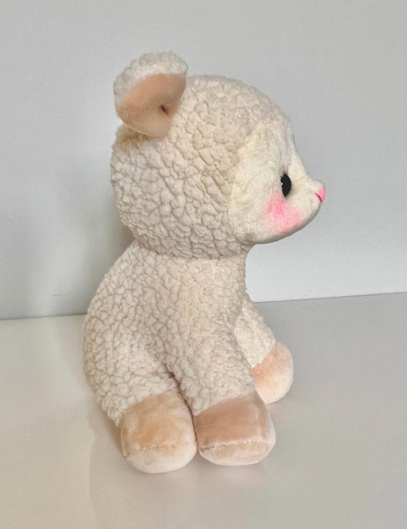 May include: A white plush lamb with a pink nose and blush on its cheeks. The lamb is sitting with its legs tucked under its body.
