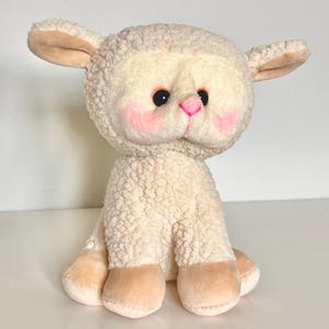 May include: A plush white lamb toy with pink cheeks and black eyes. The lamb is sitting with its legs tucked underneath it.