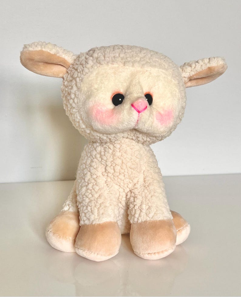 May include: A plush white lamb toy with pink cheeks and black eyes. The lamb is sitting with its legs tucked underneath it.