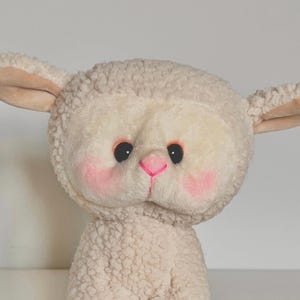 May include: A plush white lamb toy with pink cheeks and a black nose. The lamb has a fluffy white coat and big, black eyes.