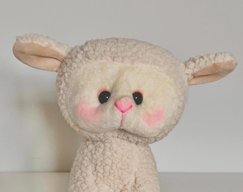 May include: A plush white lamb toy with pink cheeks and a black nose. The lamb has a fluffy white coat and big, black eyes.