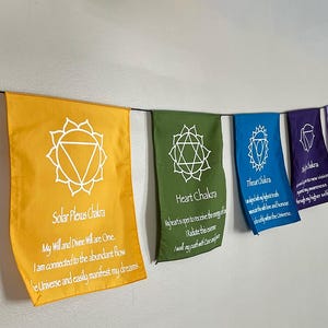 May include: A set of seven chakra flags hanging from a string. The flags are in the colors of the chakras, with white symbols and text. The text includes the name of each chakra and a short affirmation. The flags are: Root Chakra, Sacral Chakra, Solar Plexus Chakra, Heart Chakra, Throat Chakra, Third Eye Chakra, and Crown Chakra.