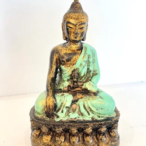 May include: A seated Buddha figurine featuring a distressed gold and turquoise finish. The figure is depicted in a meditative pose with one hand resting on the knee, set upon a decorative pedestal base with carved detailing along the bottom edge.