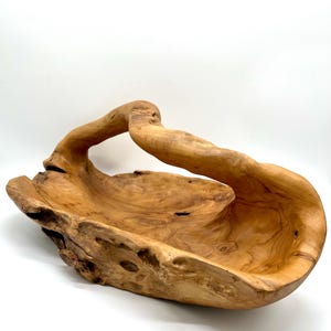 May include: A large, hand-carved wooden bowl with a natural, organic shape. The bowl is made from light brown wood and features a curved handle. The interior of the bowl is smooth, while the exterior has a rough, textured surface. A decorative piece for the home.