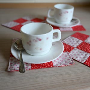 Puede incluir: Tazas y platillos blancos con diseño floral sobre posavasos de patchwork rojos y blancos. Una cuchara plateada descansa sobre un platillo. La escena se desarrolla sobre una superficie de madera clara.