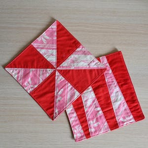 Op de afbeelding: Twee handgemaakte stoffen onderzetters. Eén is vierkant met een windmolenontwerp in rode, roze en witte bloempatronen. De andere is rechthoekig met rood-roze gestreepte secties. Beide hebben rood gestikte randen.