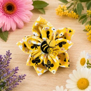 May include: A fabric flower accessory featuring a yellow background with a pattern of white daisies, small pink hearts, and black bees. The centre of the flower is adorned with a yellow and black bee-shaped embellishment with a smiling face.