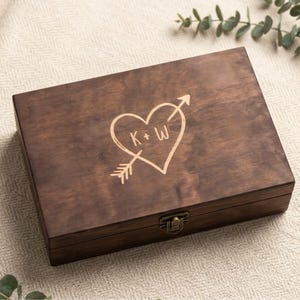 May include: A rectangular dark brown wooden box with a hinged lid and a brass-colored metal latch. The lid features a laser-engraved design of a heart pierced by an arrow, with the letters K + W centered inside the heart shape.
