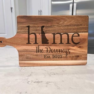 May include: A rectangular wooden cutting board with a handle. The word "home" is engraved in large letters, with the letter "o" replaced by the outline of Delaware. Below, it reads "The Downeys Est. 2023".