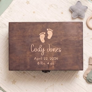 May include: A rectangular dark brown wooden keepsake box featuring an engraved design of two baby footprints, the name Cody Jones, the date April 22, 2026, and the weight 8 lbs. 4 oz. centered on the lid with a metal latch at the bottom edge.