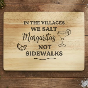 May include: Wooden cutting board with the text "IN THE VILLAGES WE SALT Margaritas NOT SIDEWALKS" in black lettering. A margarita glass and a lime slice are also illustrated on the board. The board is rectangular and sits on a wooden surface.