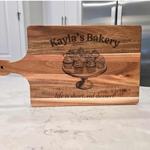 May include: Wooden cutting board with a handle, engraved with "Kayla's Bakery" and a cupcake design on a cake stand. The phrase "life is short, eat dessert" is also engraved on the board. The board is made of light brown wood.