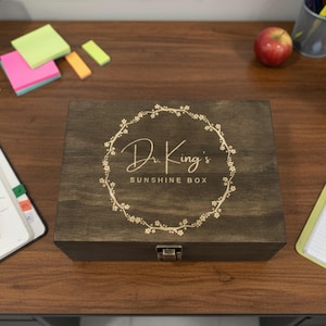 May include: A dark stained wooden box with the text "Dr. King's Sunshine Box" in a floral wreath design. The box is closed with a metal clasp. The box is on a wooden desk with office supplies.