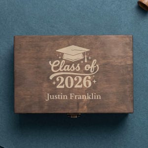 May include: A dark brown wooden box with a graduation cap graphic and the text "Class of 2026" and "Justin Franklin" engraved on the lid. The box has a small metal clasp.