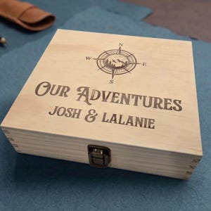 May include: A square light-colored wooden box with a metal latch closure. The lid features a laser-engraved compass rose icon above the text "OUR ADVENTURES" and the names "JOSH & LALANIE" in a bold, dark font centered on the top surface.
