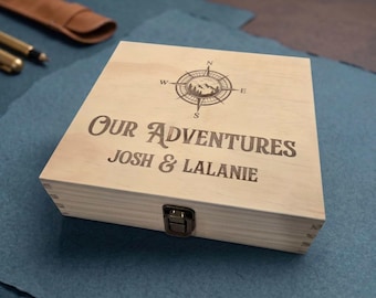 May include: A square light-colored wooden box with a metal latch closure. The lid features a laser-engraved compass rose icon above the text "OUR ADVENTURES" and the names "JOSH & LALANIE" in a bold, dark font centered on the top surface.