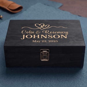 May include: A rectangular black wooden box with a metal latch. The lid features gold engraved text reading Colin & Rosemary JOHNSON, the date May 22, 2025, and a graphic of two interlocking hearts above the names. The box has a visible wood grain.