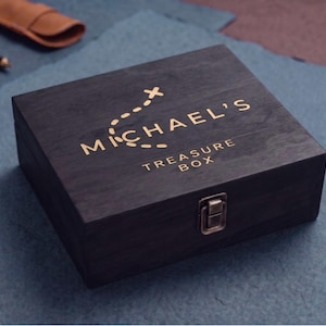 May include: A dark charcoal wooden box with a metallic latch closure. The lid features gold lettering that reads "MICHAEL'S TREASURE BOX" with a dashed path graphic above the name. The box has a visible wood grain texture and a rectangular shape.