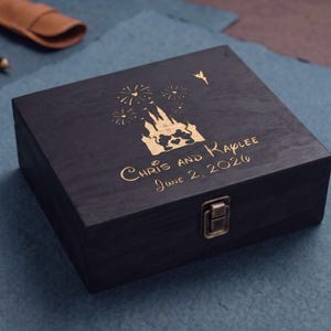 May include: A square black wooden box with a gold-colored metal latch. The lid features a gold-engraved design of a castle, fireworks, and a small fairy, along with the text "Chris and Kaylee" and the date "June 2, 2026" in a script font.