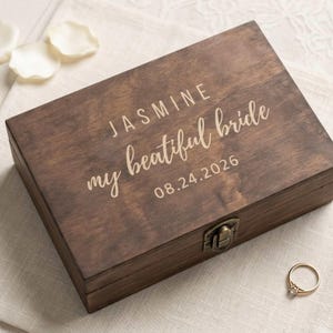 May include: A rectangular dark brown wooden box with a metal latch. The lid is engraved with the text JASMINE, my beatiful bride, and 08.24.2026. A thin gold-toned metal ring is positioned near the bottom right corner of the wooden box.