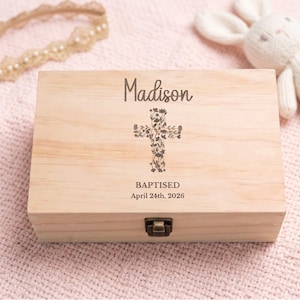 May include: A rectangular light-colored wooden keepsake box with a metal latch. The lid is engraved with the name Madison, a floral cross design, and the text BAPTISED April 24th, 2026. The box has a smooth finish and visible natural wood grain.