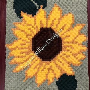 Op de afbeelding: Een haakpatroon met een zonnebloem met gele bloemblaadjes, een bruine kern en groene bladeren. Het patroon is op een groene achtergrond en bevat de tekst "August William Designs".