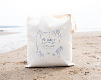 Puede incluir: Bolsa de lona blanca con texto azul e ilustraciones. El texto dice "Ansley's Last Toast on the coast" con la fecha y la ubicación debajo. Las ilustraciones incluyen conchas marinas, palmeras y copas de champán.