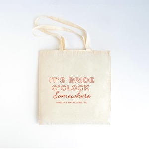 May include: Cream-colored tote bag with the text "IT'S BRIDE O'CLOCK Somewhere" in red. The text "AMELIA'S BACHELORETTE" is printed below. The bag has two cream-colored handles.