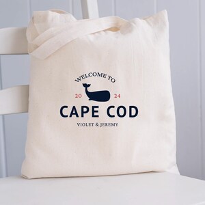 May include: A white canvas tote bag with a navy blue whale logo and the text "Welcome to Cape Cod 2024 Violet & Jeremy".