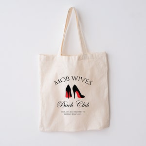 K&ouml;nnte beinhalten: Eine wei&szlig;e Canvas-Tragetasche mit schwarzem Text, der "MOB WIVES Bach Club" lautet, und einer Grafik von zwei schwarzen High Heels mit roten Sohlen. Der Text unter der Grafik lautet "VIOLET'S BACHELORETTE MIAMI BEACH FL."