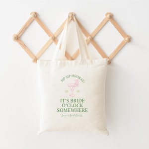May include: Off-white canvas tote bag with the text "SIP SIP HOORAY!" and "IT'S BRIDE O'CLOCK SOMEWHERE" in green and pink. A cocktail graphic is centered above the text. The bag is hanging on a wooden wall rack.