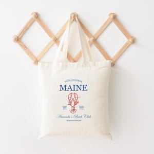 May include: A white canvas tote bag with blue and red text and a red lobster illustration. The text reads "With love from Maine, Est. 2004, Amanda's Beach Club, Kennebunkport".