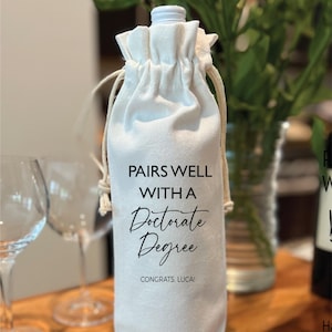 May include: A white wine bottle bag with a drawstring closure. The bag features the text "PAIRS WELL WITH A Doctorate Degree" in black script, with "CONGRATS, LUCA!" below. The bag is on a wooden surface.