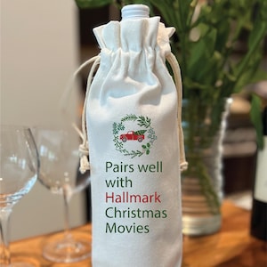 May include: White wine bottle bag with a red truck carrying a Christmas tree in a green wreath. The bag has the text "Pairs well with Hallmark Christmas Movies".