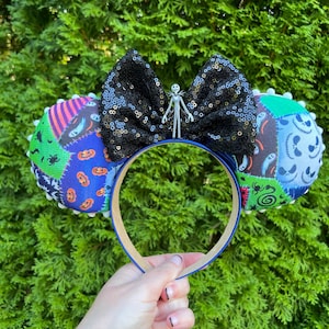 May include: A Halloween-themed headband with padded ears featuring patchwork patterns and a black sequined bow. A small Jack Skellington figure is centered on the bow. The headband is held against a backdrop of green foliage.